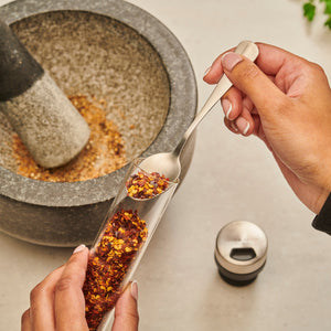 A person spoons dried chili flakes from the Cole & Mason Shadwell Drawer-Fit Spice Storage into a stone mortar, with a pestle and spice lid nearby on a light countertop.