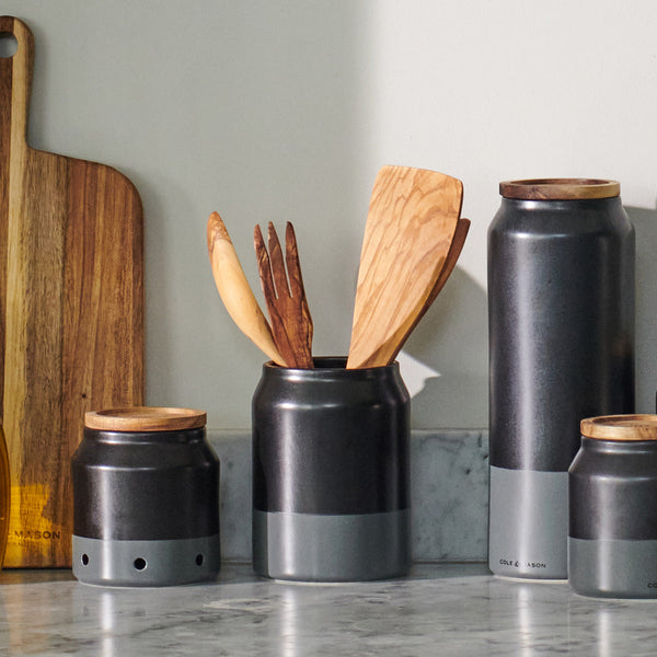 The Cole & Mason Linton Ceramic Utensil Pot holds cooking tools on a marble countertop beside a wooden cutting board, set against a light-colored wall.