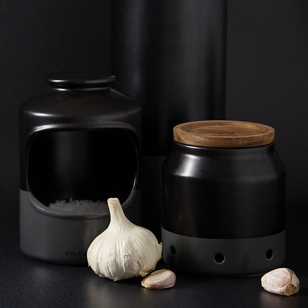 The Cole & Mason Strethall Ceramic Salt Pig, a black ceramic salt keeper with an open front and matching wooden-lidded jar, is arranged beside a garlic bulb on a dark surface against a dark background.