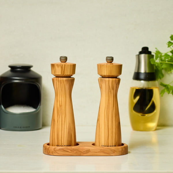 #salt&pepperset The Cole & Mason Portobello Olive Wood Salt & Pepper Mills stand on a matching tray, each with a precision grinding mechanism. In the background are a black salt container, an oil bottle, and fresh herbs on a light kitchen counter.
