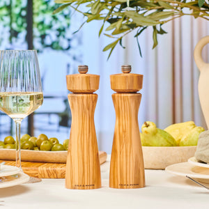 #pepper The Cole & Mason Portobello Olive Wood Salt & Pepper Mills with adjustable grind rest on a dining table set with a glass of white wine, green olives, and fruit, sunlight streaming through large windows in the background.
