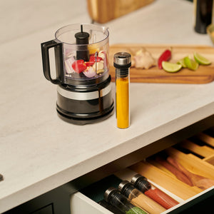 A food processor, yellow spice tube, and a cutting board with lime, ginger, and apple slices rest on a counter above the Cole & Mason Shadwell Drawer-Fit Spice Storage.