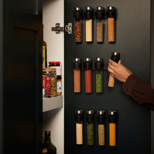 A person chooses a spice jar from the Shadwell Slim-Mount Spice Storage by Cole & Mason, a wall-mounted rack with 12 magnetic containers on a dark pantry door, efficiently organizing herbs and spices.
