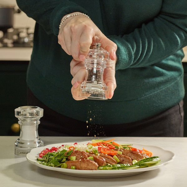 A person in a green sweater grinds pepper onto a plate of food with vegetables and meat, using the Cole & Mason Acrylic Pepper Mill from the 104 mm Salt Shaker & Pepper Mill Set. Another grinder from the set rests on the table nearby, next to the matching acrylic salt shaker.