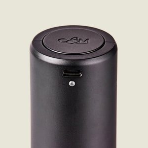 Close-up of the Cole & Mason Brixton Electric Mill Matt Black GS, a cylindrical black electronic device featuring a USB-C port and a small indicator light. Designed for one-handed operation, this electric mill has an embossed C&M logo on its circular lid and contains a durable ceramic grinder inside.