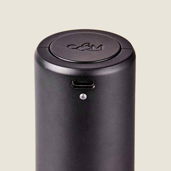 Close-up of the Cole & Mason Brixton Electric Mill Matt Black GS, a cylindrical black electronic device featuring a USB-C port and a small indicator light. Designed for one-handed operation, this electric mill has an embossed C&M logo on its circular lid and contains a durable ceramic grinder inside.