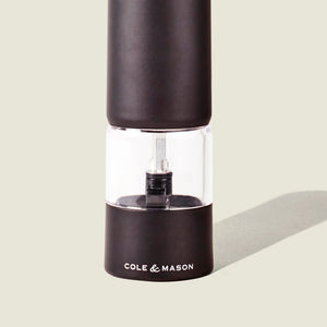 A black and clear Cole & Mason Brixton Electric Mill Matt Black GS pepper grinder with a ceramic mechanism stands upright on a light background, casting a faint shadow to the right.