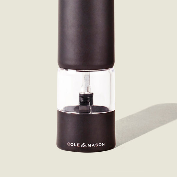 A black and clear Cole & Mason Brixton Electric Mill Matt Black GS pepper grinder with a ceramic mechanism stands upright on a light background, casting a faint shadow to the right.