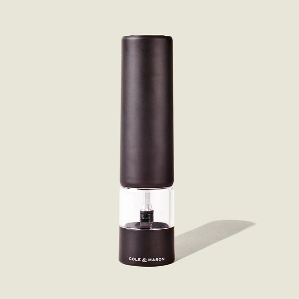 The Cole & Mason Brixton Electric Mill Matt Black GS is an electric pepper grinder with a ceramic grinding mechanism and a clear middle section for easy viewing, designed for convenient one-hand operation. The mill, branded with the Cole & Mason logo on its base, stands upright against a light beige background.