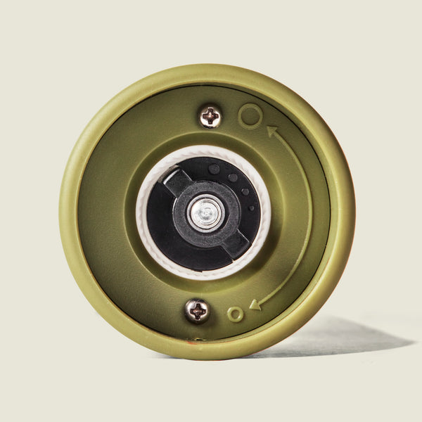 Close-up of the bottom of the Cole & Mason Brixton Electric Mill Olive Green GS, highlighting its green cylindrical design, visible screws, a rotating mechanism with a ceramic grinder, and directional arrows for convenient one-handed operation.
