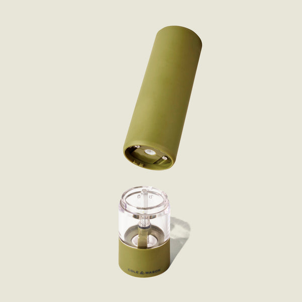 A Cole & Mason Brixton Electric Mill Olive Green GS, with its cylindrical top section removed to display the ceramic grinding mechanism and clear base featuring the Cole & Mason logo, is set against a light beige background—engineered for effortless one-handed operation.