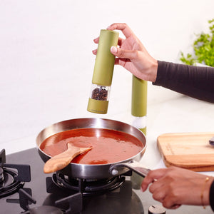 Using the Cole & Mason Brixton Electric Mill Olive Green GS with a durable ceramic grinder, someone effortlessly seasons a pan of red sauce simmering on the stovetop, operating the mill one-handed while a wooden spoon rests in the sauce and a cutting board sits in the background. This design makes seasoning quick and easy.