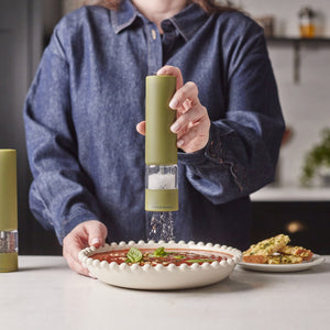 A person in a blue shirt grinds pepper over a bowl of soup garnished with mint using the Cole & Mason Brixton Electric Mill Olive Green GS. Slices of bread rest on a plate in the background on a white kitchen counter.