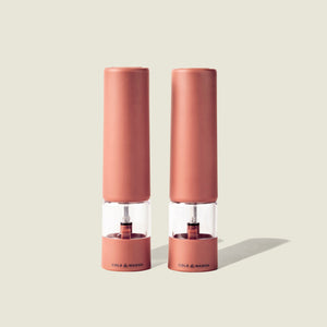 Two identical Cole & Mason Brixton Electric Mill Terracotta GS grinders stand side by side on a light beige background, casting soft shadows to the right. Featuring a durable ceramic grinding system and convenient USB-C charging, these mills combine modern functionality with elegant design.