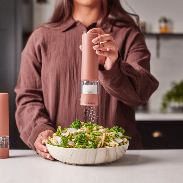 A person in a brown shirt uses the Cole & Mason Brixton Electric Mill Terracotta GS, featuring a ceramic grinding system, to season a bowl of pasta with vegetables on a white kitchen counter.