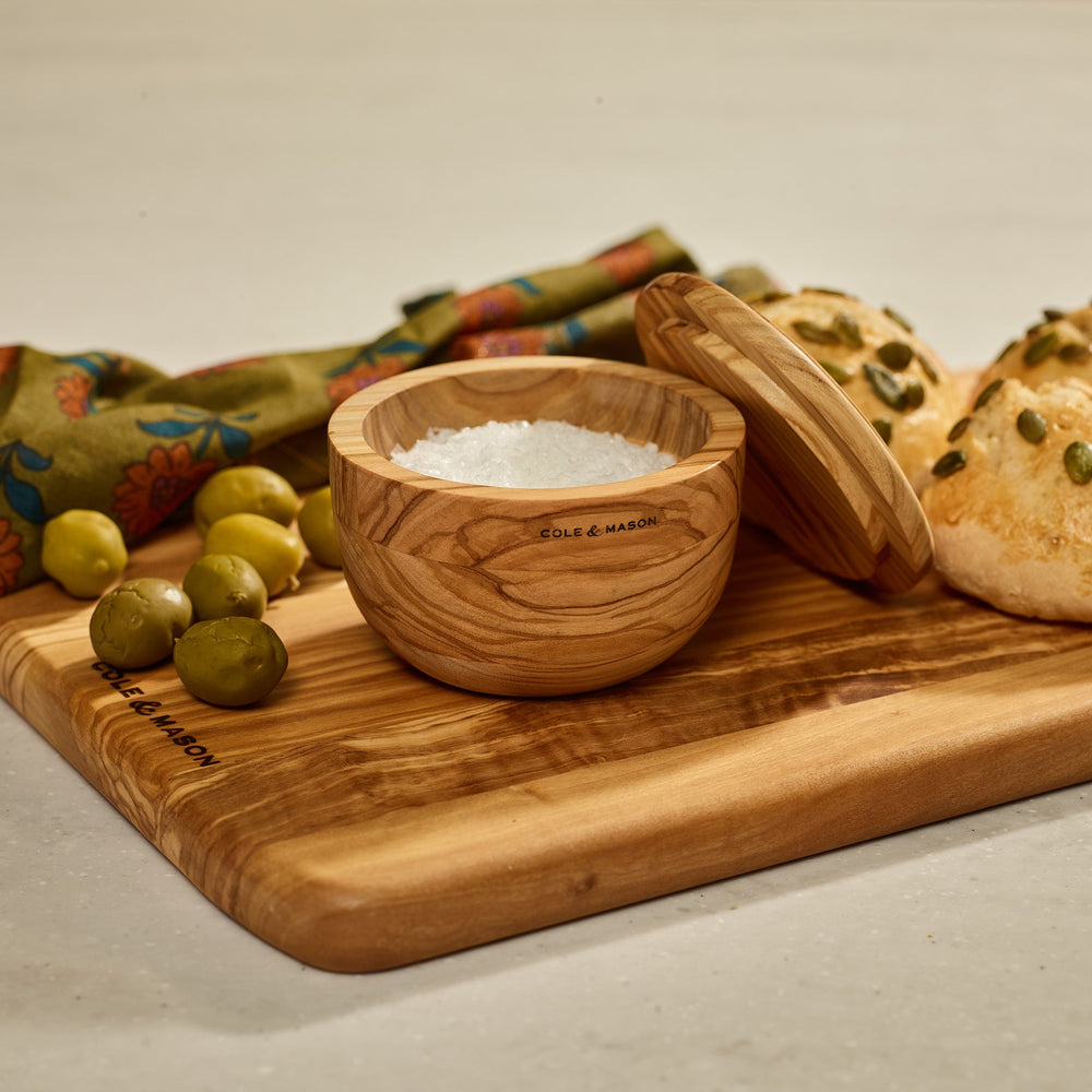The Portobello Olive Wood Salt Cellar by Cole & Mason features natural grain patterns, a matching lid, and the brand logo on the front, adding timeless charm to your kitchen decor. Shown against a plain light background.