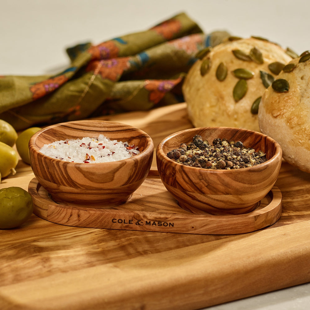 The Cole & Mason Portobello Olive Wood Pinch Pot Set features two small pots with unique grain patterns on a matching tray engraved with the brand name. The set is displayed against a plain, light beige background.