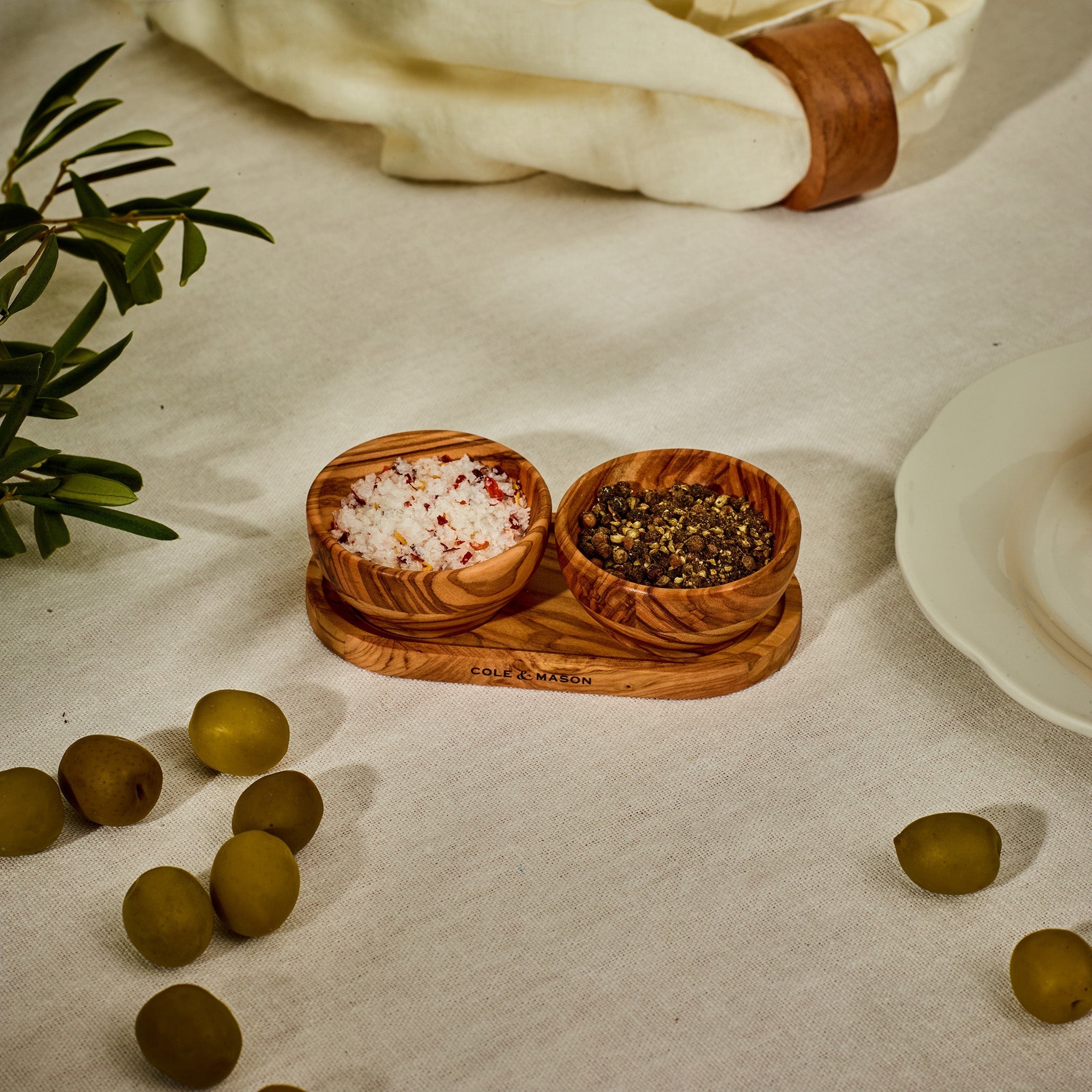 The Cole & Mason Portobello Olive Wood Pinch Pot Set features two uniquely grained pots on a seasoning tray, perfect for salt and pepper. Shown on a white cloth next to green olives, a plate, and a napkin with a leather ring.