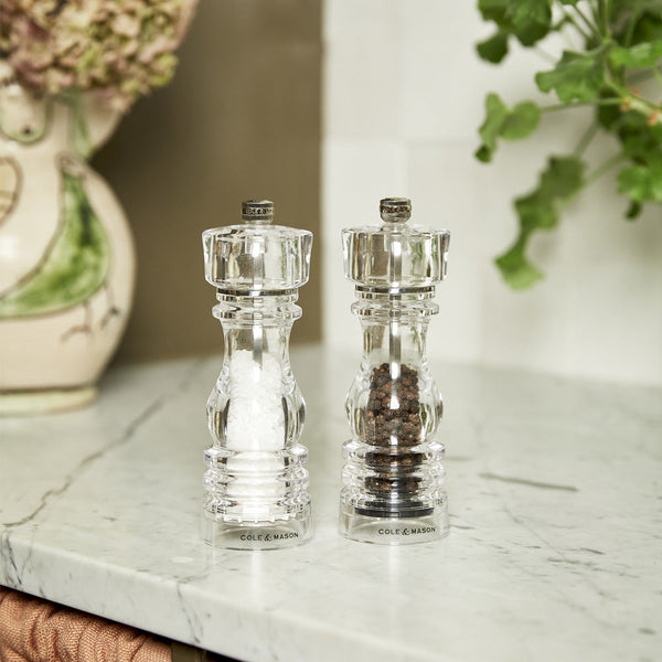 Two clear Cole & Mason mills, one with white sea salt and one with black peppercorns, sit side by side on a marble countertop in a kitchen with plants and decorative items in the background.