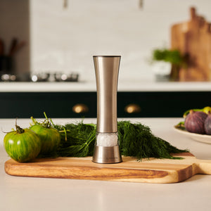 The Cole & Mason Burford Electronic Single Mill, made of stainless steel with adjustable grind settings, rests on a wooden cutting board next to fresh herbs, green tomatoes, and figs in a modern kitchen.