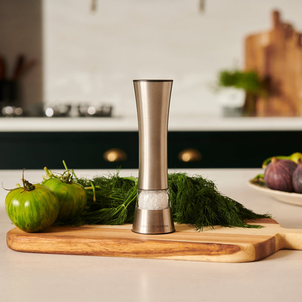 The Cole & Mason Burford Electronic Single Mill, made of stainless steel with adjustable grind settings, rests on a wooden cutting board next to fresh herbs, green tomatoes, and figs in a modern kitchen.