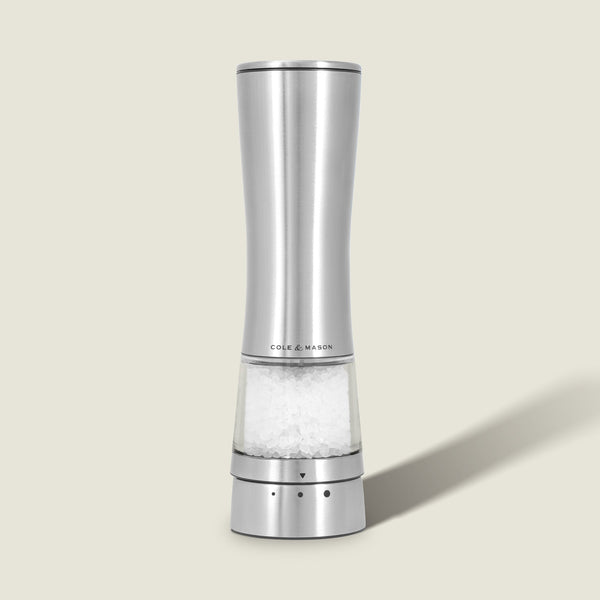 A Cole & Mason Electric Derwent Salt & Pepper Mill, crafted from stainless steel with a transparent base filled with coarse salt, stands upright on a light beige background.