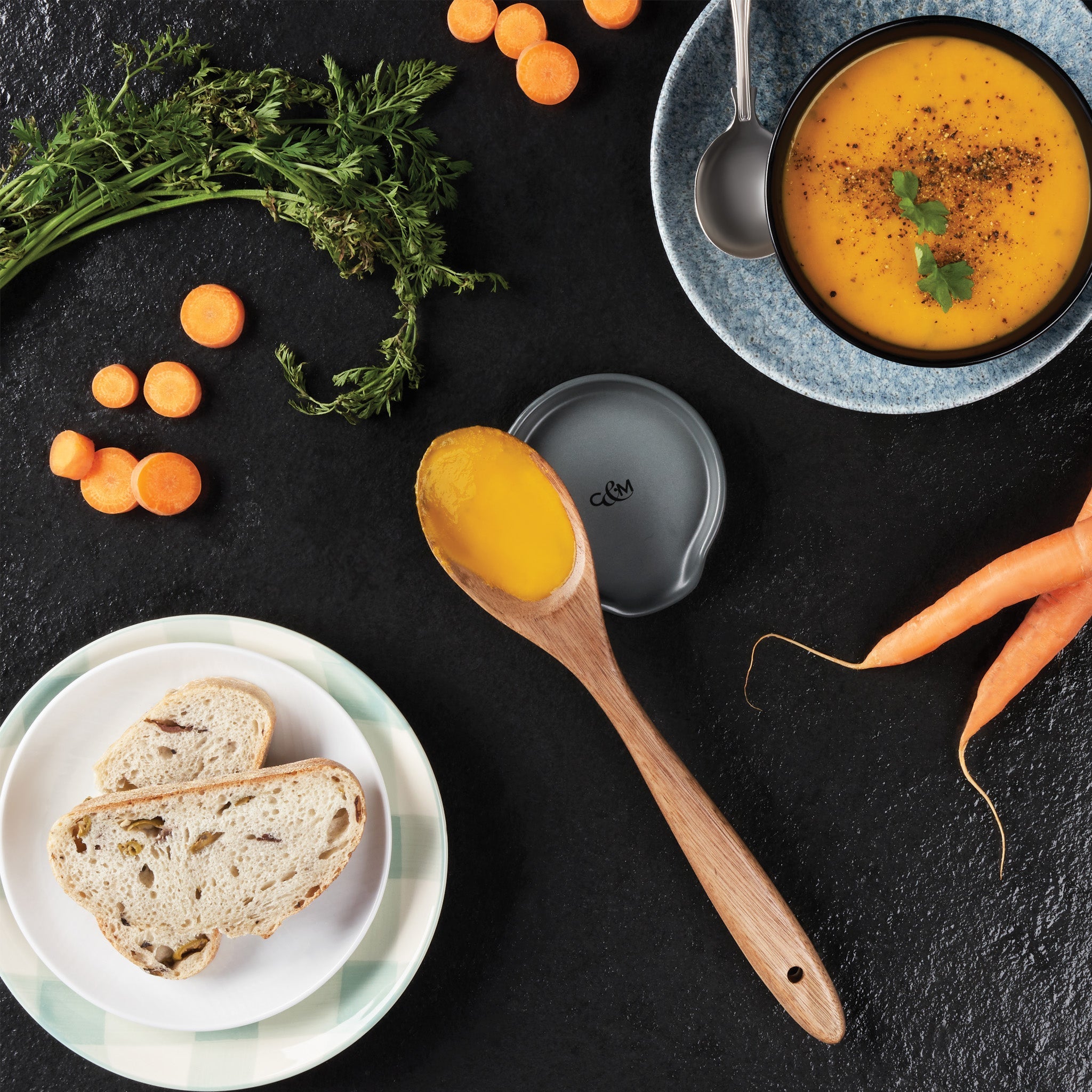 Overhead view of a wooden spoon filled with carrot soup, a bowl of soup with cilantro, sliced carrots, carrot tops, and two slices of bread on a plate, all arranged on a black surface.