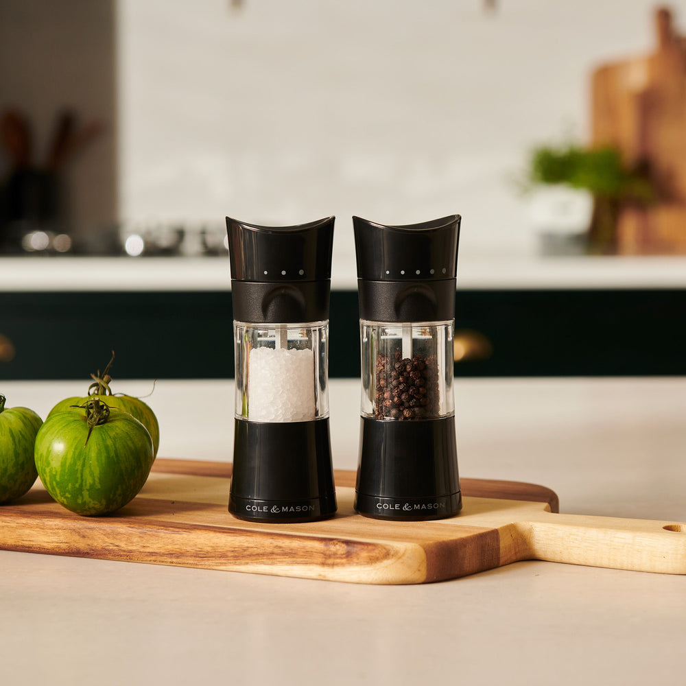 The Cole & Mason Harrogate Salt & Pepper Mill Set features stylish grinders with black accents and adjustable settings—one filled with coarse salt, the other with whole peppercorns—both upright on a plain light background.