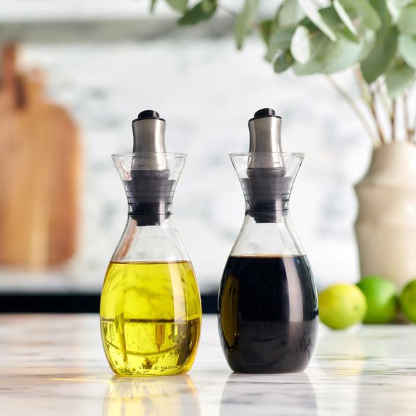 Two Cole & Mason Haverhill Oil & Vinegar Pourers, one with golden olive oil and the other with dark balsamic vinegar, rest on a marble countertop. Blurred kitchen items and green limes complete the welcoming atmosphere.