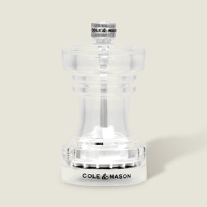 Showcased against a light beige background, the Cole & Mason Acrylic Salt Shaker & Pepper Mill Set (104 mm) features a clear acrylic body with a silver knob on top and “COLE & MASON” inscribed in black on the base; this elegant pepper mill offers an adjustable grinder for customized seasoning.