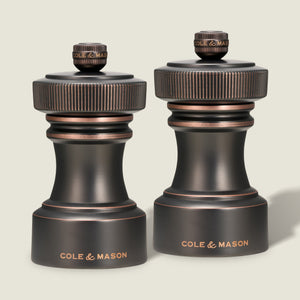 Two Cole & Mason Hoxton Salt and Pepper Mills in antique bronze, each with a dark finish, textured knurled tops, and the Cole & Mason brand name engraved in gold at the base, stand side by side against a plain light background. This sophisticated set features a precision grinding system for effortless seasoning every time.