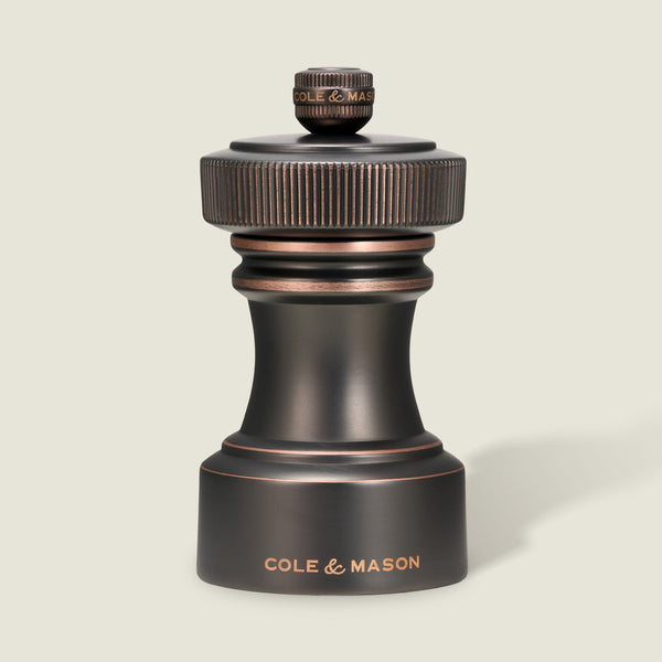 A Cole & Mason Hoxton Salt and Pepper Mill, showcasing an antique bronze finish with a ridged top and engraved logo, rests on a light background. This elegant piece features a precision grinding system, making it the perfect addition to any salt and pepper mill set.
