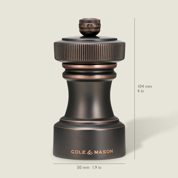 A dark metallic pepper grinder from the Cole & Mason Hoxton Salt and Pepper Mill Set stands upright, showcasing an antique bronze finish. Dimensions are indicated as 104 mm (4 in) tall and 50 mm (1.9 in) wide, set against a light beige background.