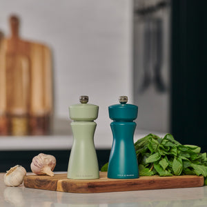 Two Cole & Mason Kenton Sage Salt & Pepper Mills, with durable ceramic mechanisms and light green and teal finishes, rest on a wooden board alongside fresh herbs and garlic, adding style to your kitchen counter.