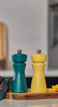 The Cole & Mason Kenton Sea Salt Blue Salt & Pepper Mill sits on a wooden board with sliced yellow squash and a patterned cloth, bringing a pop of style and featuring a high-performance ceramic mechanism for contemporary kitchens.