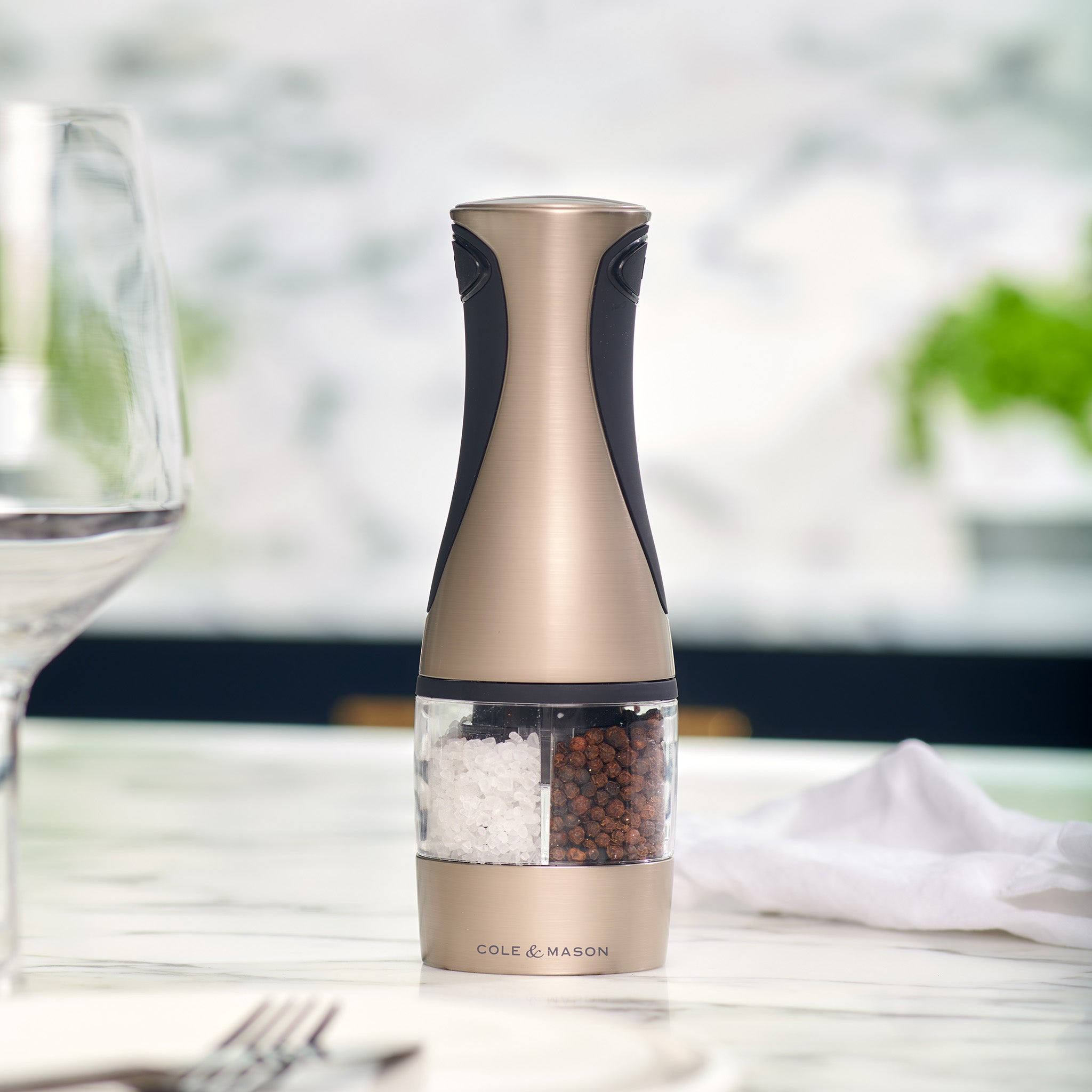 A metallic Cole & Mason Kew Electronic 2-in-1 Duo Mill salt and pepper grinder sits on a marble table near a wine glass and white napkin, with a blurred green plant in the background.