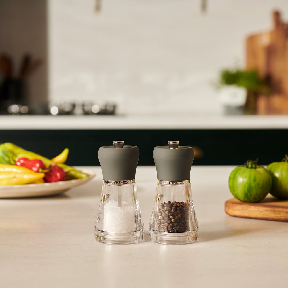 The Cole & Mason Napoli Salt & Pepper Mill Set features clear glass bodies with gray tops, elegant design, and premium grinders. Filled with white salt crystals and whole brown peppercorns, they stand side by side on a light background.