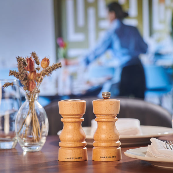 A Cole & Mason London Natural Beech Salt Shaker & Pepper Mill Set rests on a restaurant table with plates, napkins, and dried flowers in a glass vase, while a blurred figure works in the background.