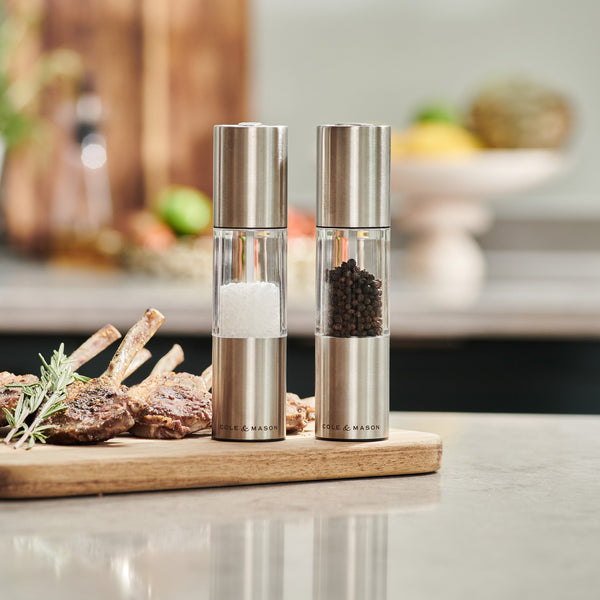 The Cole & Mason Oslo Salt & Pepper Mill Set, featuring adjustable grinding mechanisms, sits on a kitchen counter beside cooked meat on a wooden board. A softly blurred background highlights precise seasoning control.