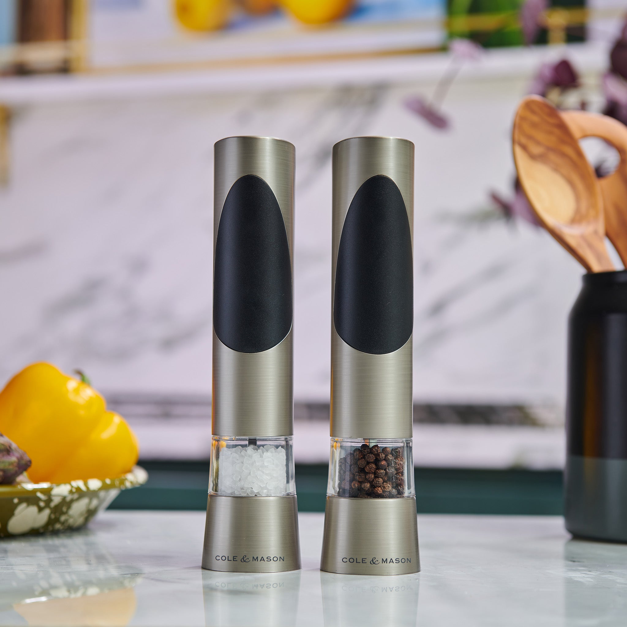 The Cole & Mason Richmond Electronic Salt & Pepper Mill Set features a sleek stainless steel and black design with adjustable grind levels, shown side by side on a kitchen counter with a yellow bell pepper and utensils in the background.