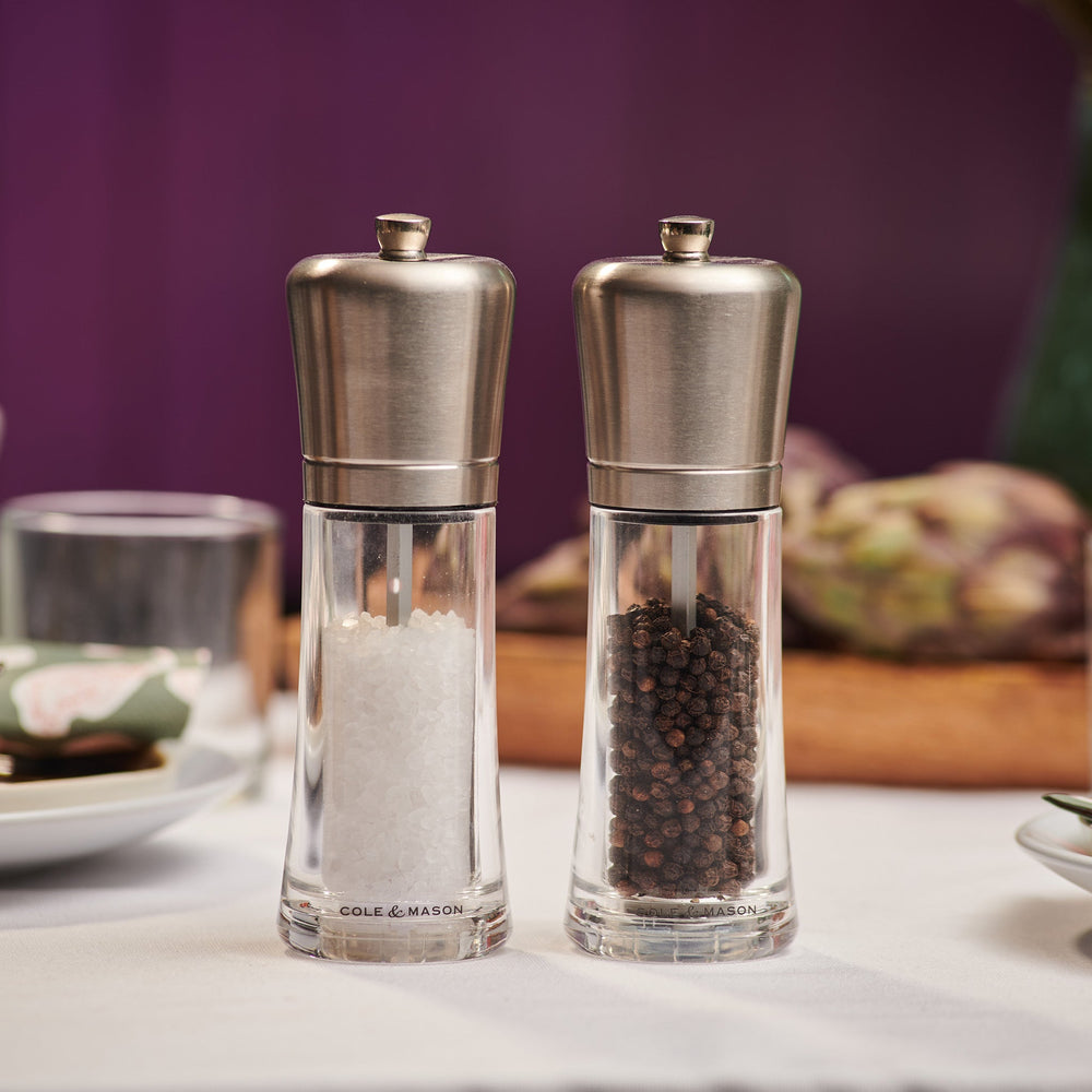 The Cole & Mason Sandown Salt & Pepper Mill Set features clear glass mills with stainless steel tops, filled with black peppercorns and coarse salt, and offers adjustable grinding for perfect seasoning control.