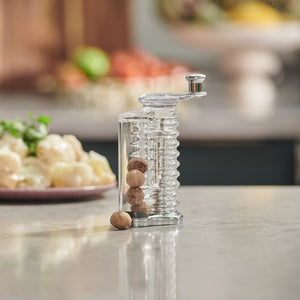 The Cole & Mason Wallis Professional Nutmeg Grinder, featuring a clear acrylic spiral design and stainless steel blade, holds whole nutmegs on a countertop with a blurred kitchen background of vegetables and fruit.