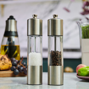 The Cole & Mason Everyday Stainless Steel Salt & Pepper Mill Set with ergonomic design and adjustable coarseness holds coarse salt and black peppercorns side by side on a kitchen counter, with blurred kitchen items in the background.