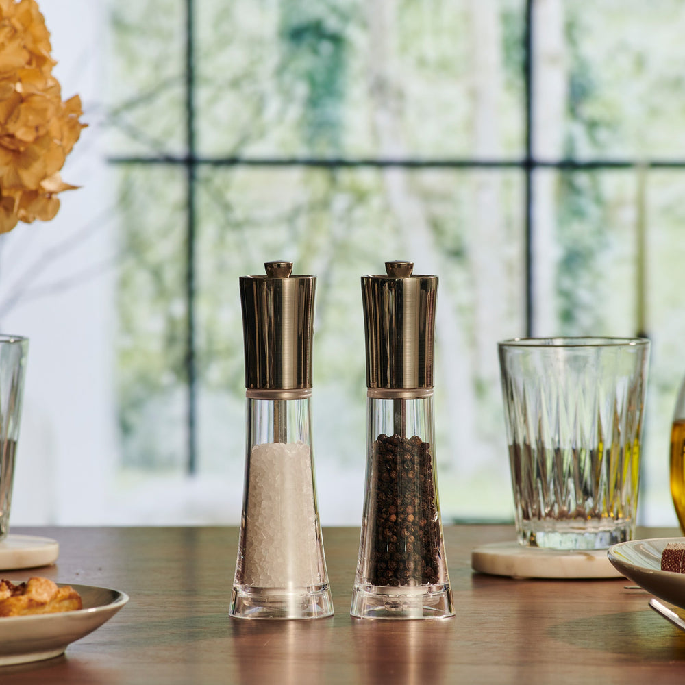 The Cole & Mason Everyday Style Salt & Pepper Mill Set includes two clear glass grinders with stainless steel tops and adjustable settings—ideal for any kitchen. One holds salt, the other peppercorns, displayed side by side on a light background.